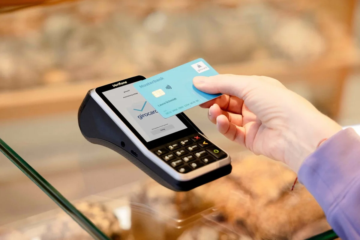 A customer tapping a turquoise girocard against a Verifone card terminal on a shop counter, the screen showing girocard contactless approval